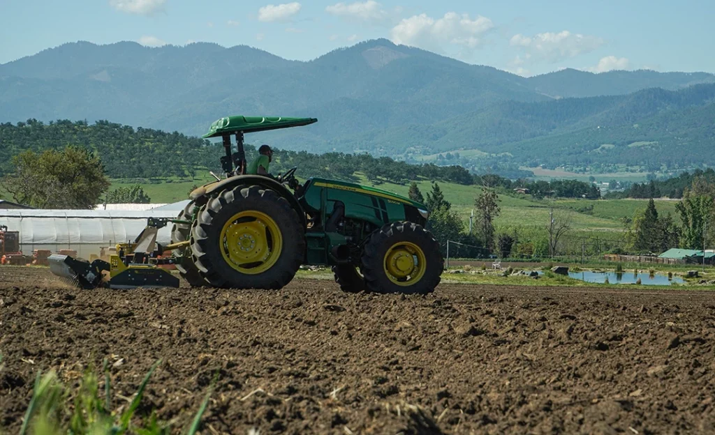 Tractor tilling the fields at fern valley farms with greenhouse, pond, and mountains in background